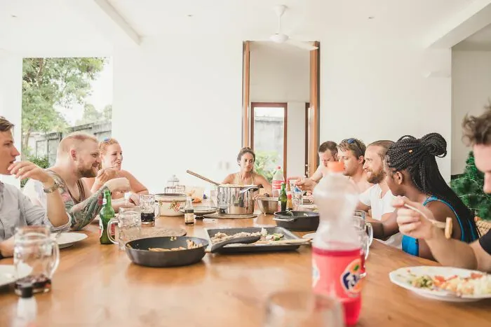 A diverse group of locals and foreigners shares a meal at a long table, highlighting potential cultural misunderstandings.