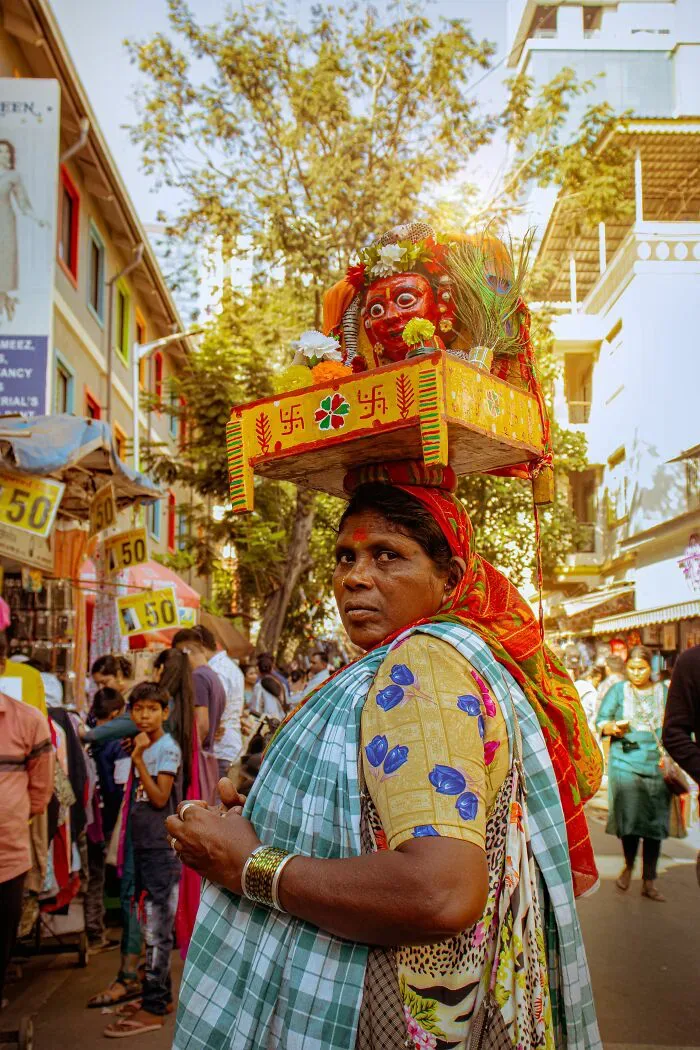 A local woman carries a vibrant religious offering on her head in a bustling market, a scene illustrating misunderstandings between locals and foreigners.