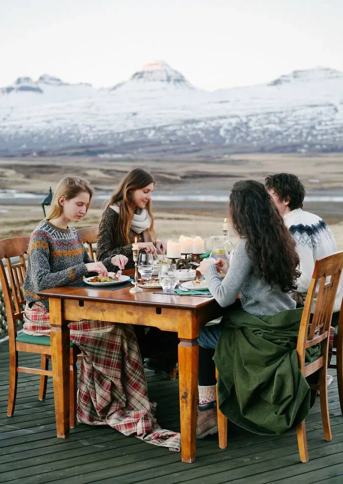 Four individuals, a mix of locals and foreigners, dine outdoors with snowy mountains in the background, a setting for cultural misunderstandings.