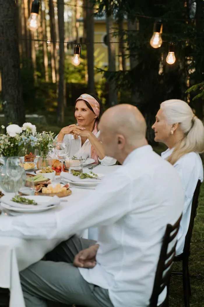 Three elderly people at an outdoor dinner table with string lights, showing interactions between locals and foreigners.