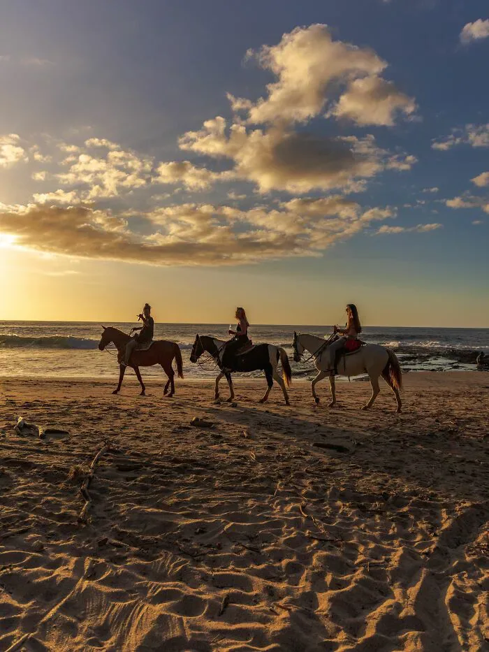 Three individuals riding horses on a beach at sunset, showing interaction between locals and foreigners.