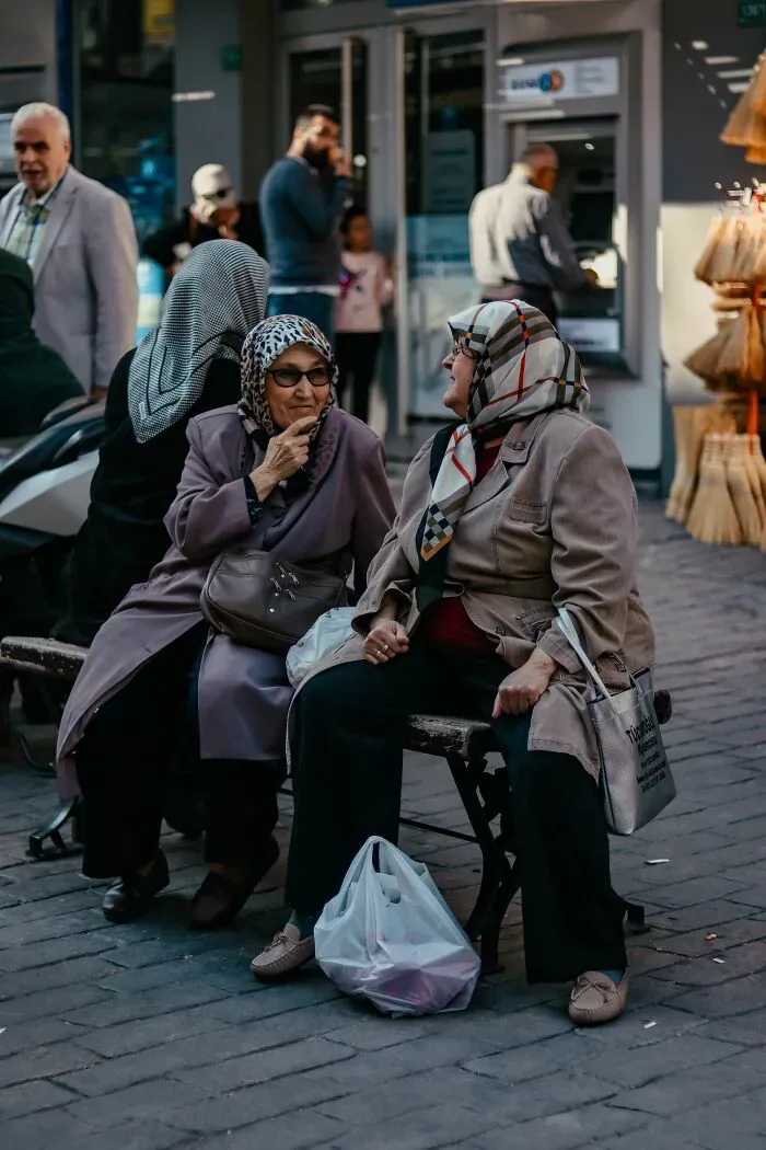 Two elderly women in headscarves sitting and conversing on a street bench, depicting interactions among locals and foreigners.