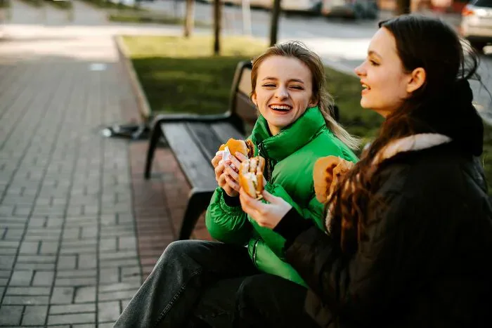 Two happy women eating burgers on a bench, highlighting interactions between locals and foreigners.