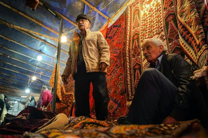 Two older men stand among tapestries in a bustling market, highlighting local and foreigner interactions.
